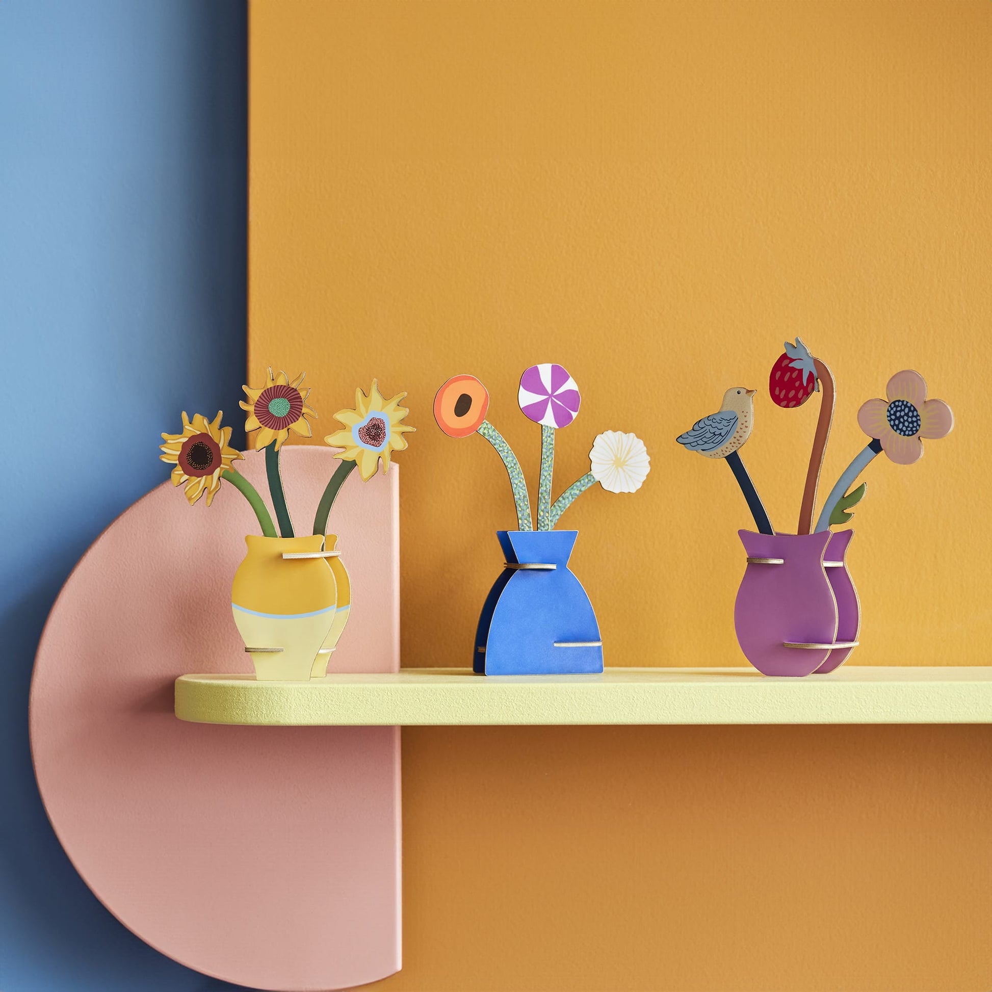 Colorful paper flower arrangements in vases on a shelf against a multicolored wall.