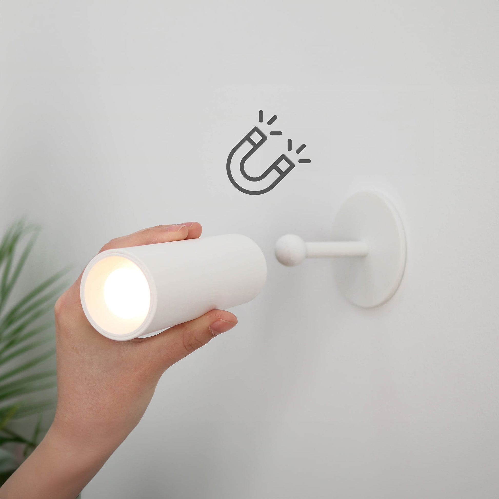Hand holding a white lamp next to a wall-mounted light fixture with a magnetic icon.