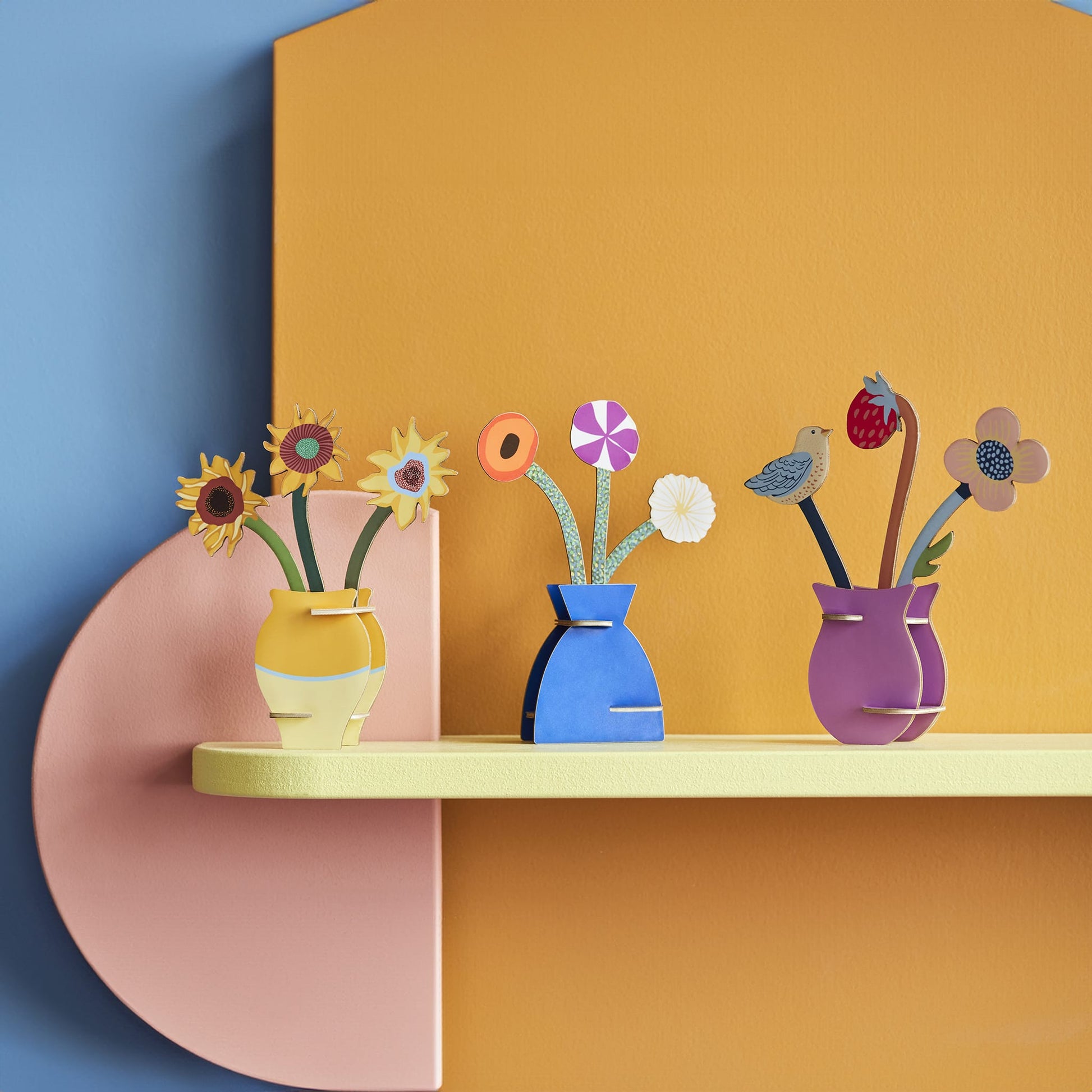 Colorful paper flowers in vases on a shelf against a multicolored wall.